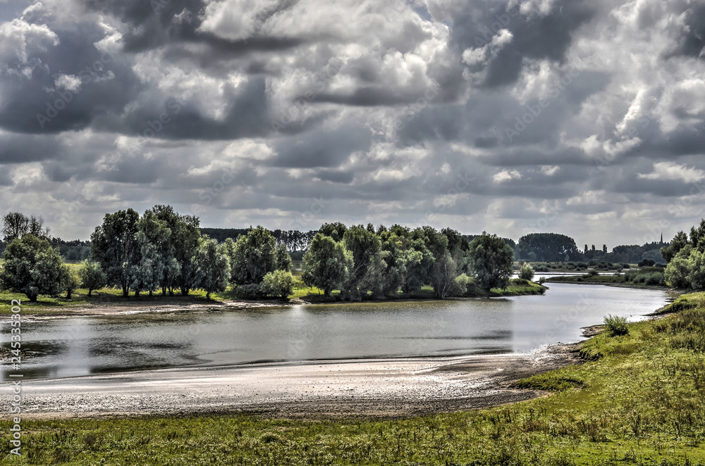 Dramatic sky over a secondary channel of the river Waal, near Tiel, The Netherlands, with sandy beaches, grassy meadows and group of trees and bushes