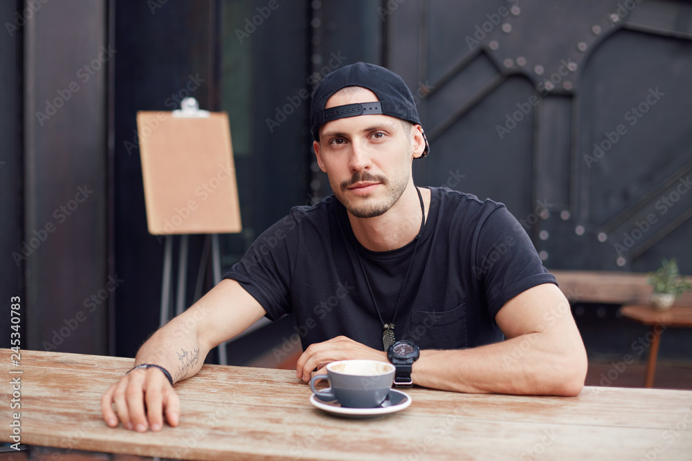 People, lifestyle concept. Handsome confident young Caucasian bearded freelancer in black t-shirt and hat, enjoying coffee break while relaxing after having finished art project, looking at camera. People, lifestyle concept. Handsome confident young Caucasian bearded freelancer in black t-shirt and hat, enjoying coffee break while relaxing after having finished art project, looking at camera.
