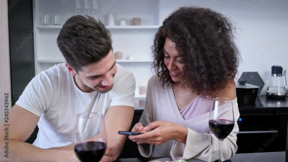 Playful loving multiethnic couple drinking wine and talking while sitting on kitchen