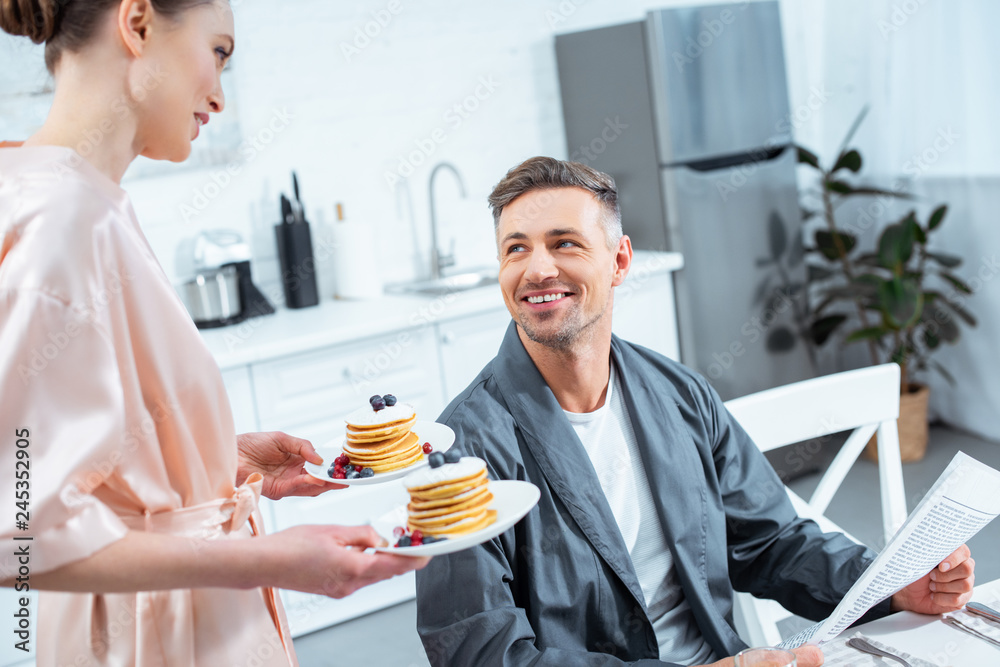 woman holding plates with pancakes while smiling man reading newspaper during breakfast in kitchen woman holding plates with pancakes while smiling man reading newspaper during breakfast in kitchen