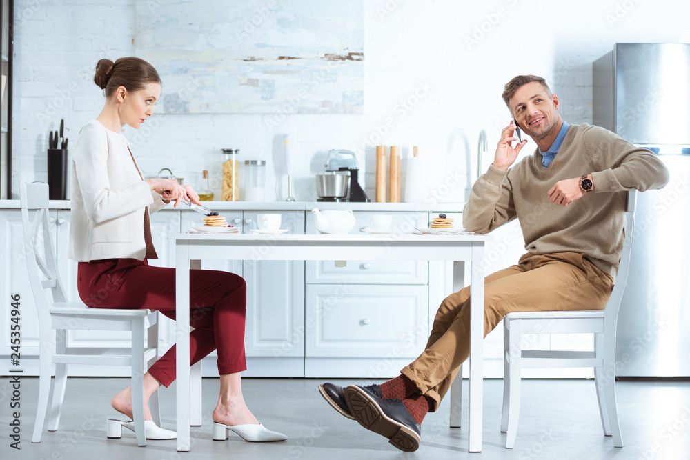 dissatisfied woman cutting pancakes while man talking on smartphone during breakfast in kitchen