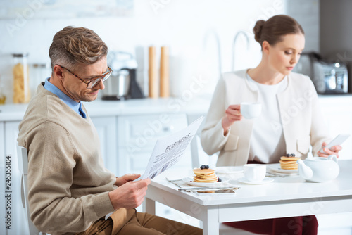 adult couple reading newspaper, drinking tea and ignoring each other during breakfast in morning