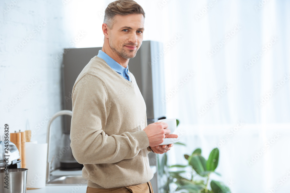 handsome man in casual clothes holding coffee cup and looking at camera in kitchen