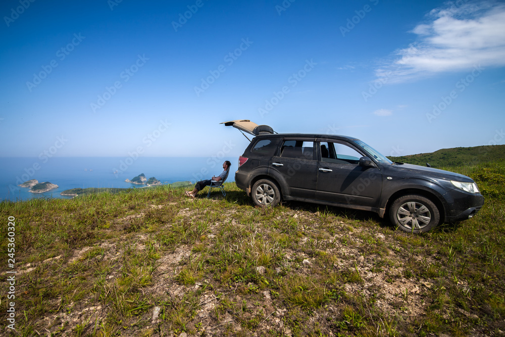Man sitting at car's shadow at the top of the hill at sea coast