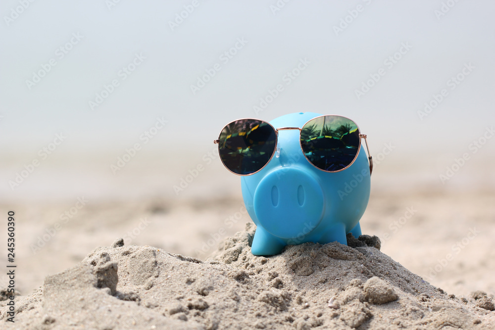 Obraz premium Blue piggy bank with sunglasses on sea beach, Saving planning for Travel budget of holiday concept