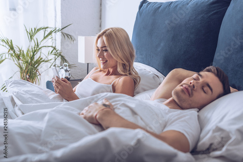 handsome young man sleeping and beautiful smiling woman using smartphone in bed