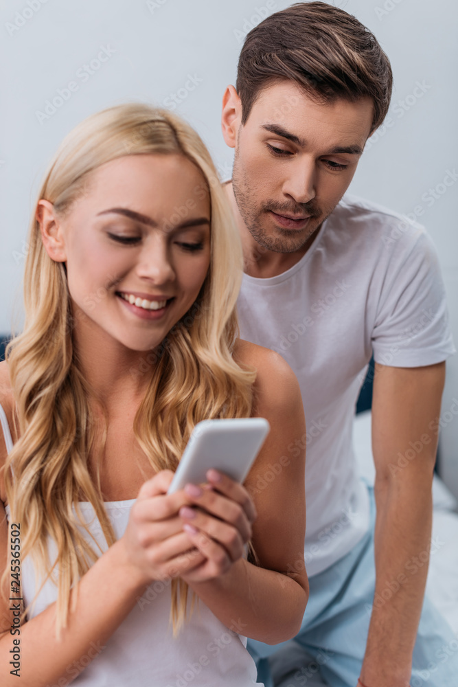 jealous young man looking at beautiful smiling wife using smartphone in bedroom jealous young man looking at beautiful smiling wife using smartphone in bedroom
