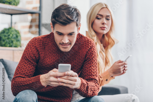 Bild auf Leinwand jealous young woman with smartphone looking at smiling boyfriend using smartphon