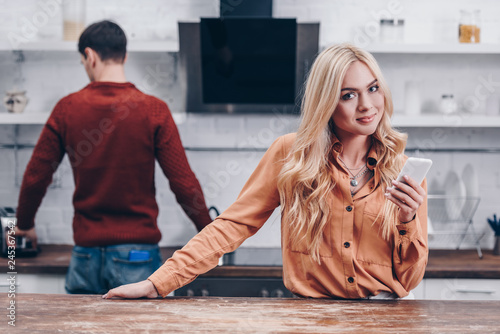 beautiful young woman using smartphone and smiling at camera while husband standing in kitchen behind photo