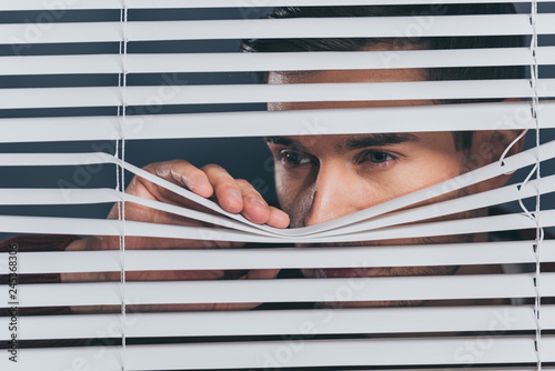 suspicious young man looking away and peeking through blinds photo