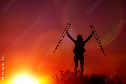 a woman on top of a mountain with medical crutches in her hands against the evening sky. achievement, conquest of the top. © gerasimov174