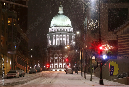 Snowy storm in a city background. Weather alert concept. Blizzard night downtown cityscape with glowing capitol building and baked cars along street. Midwest USA, Wisconsin, Madison.