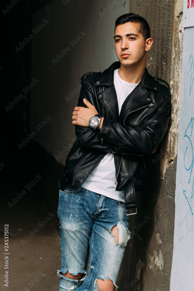 young, handsome guy in a white T-shirt, black, leather jacket and blue, torn jeans is on the street, model, city walk, live style. in white sneakers