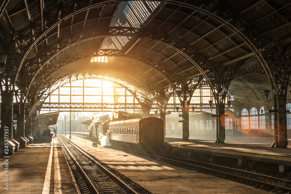Fototapeta premium Stara stacja kolejowa z pociągiem i lokomotywą na peronie oczekującym na odjazd. Promienie słońca wieczorem w łukach dymu.