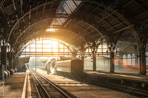 Old railway station with a train and a locomotive on the platform awaiting departure. Evening sunshine rays in smoke arches.