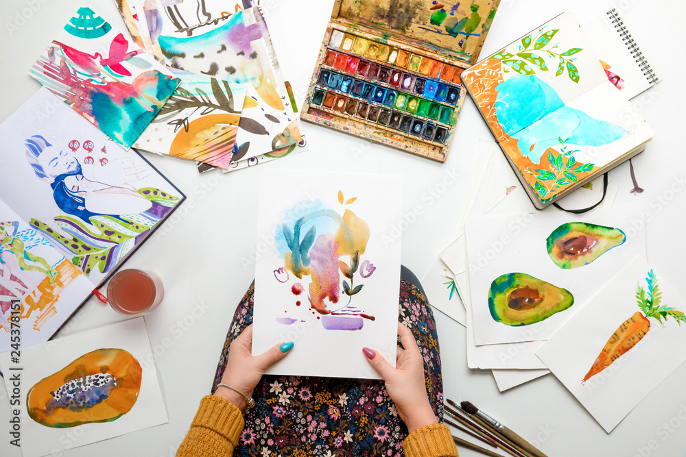 Fototapeta premium top view of woman holding watercolor drawing while surrounded by color pictures