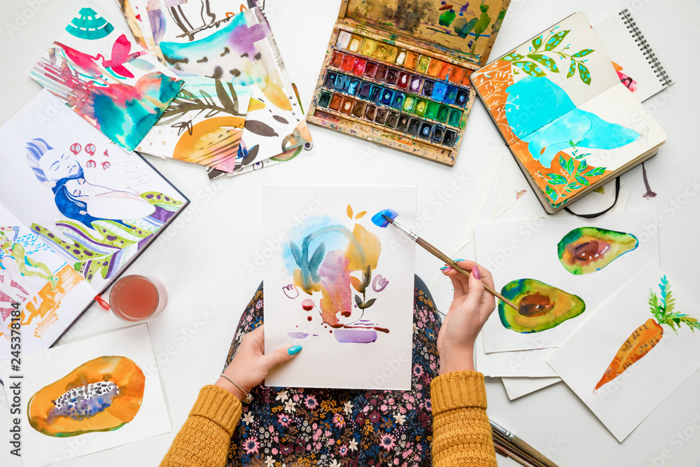Obraz premium top view of woman holding drawing on knees and painting in it with watercolors paints while surrounded by colored pictures