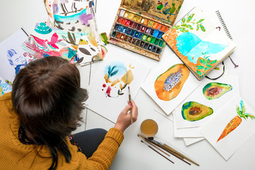 top view of woman painting with watercolors paints while surrounded by color drawings and drawing utensils