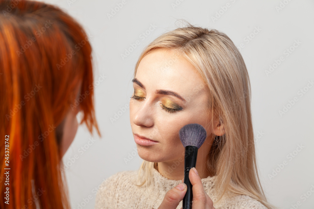 Makeup artist girl with red hair puts make-up on a blonde model with eyes closed, holds a brush in her hands and puts contouring of her face on a white background in visage studio. Makeup artist girl with red hair puts make-up on a blonde model with eyes closed, holds a brush in her hands and puts contouring of her face on a white background in visage studio.