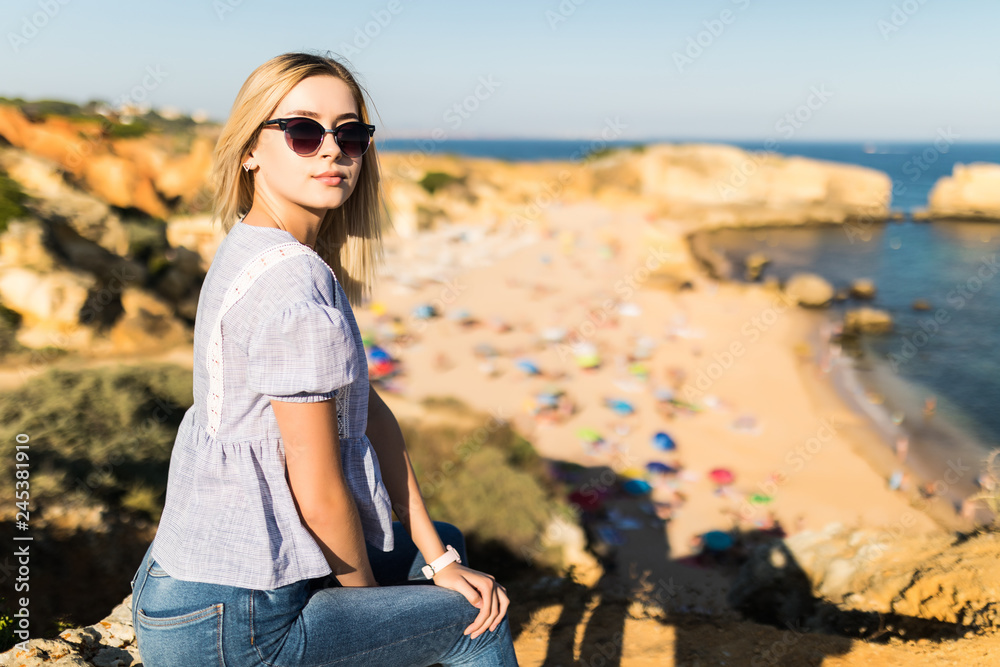 Young pretty woman wear in sunglasses and wear casual clothes sitting on hight stone and enjoying the view on the beach