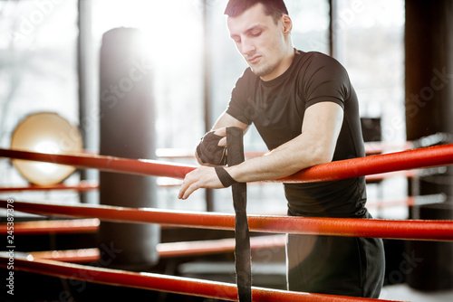 Portrait of a young athletic man winding bandage on the wrists, preparing for training on the boxing ring at the gym