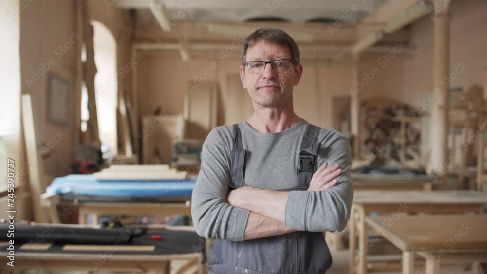 Mature woodworker in eyeglasses and overalls approaching camera and standing cross-armed in his workshop