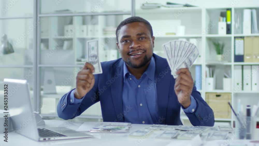 Excited african american businessman holding dollar banknotes with both hands, smiling and telling something at camera in the office Excited african american businessman holding dollar banknotes with both hands, smiling and telling something at camera in the office