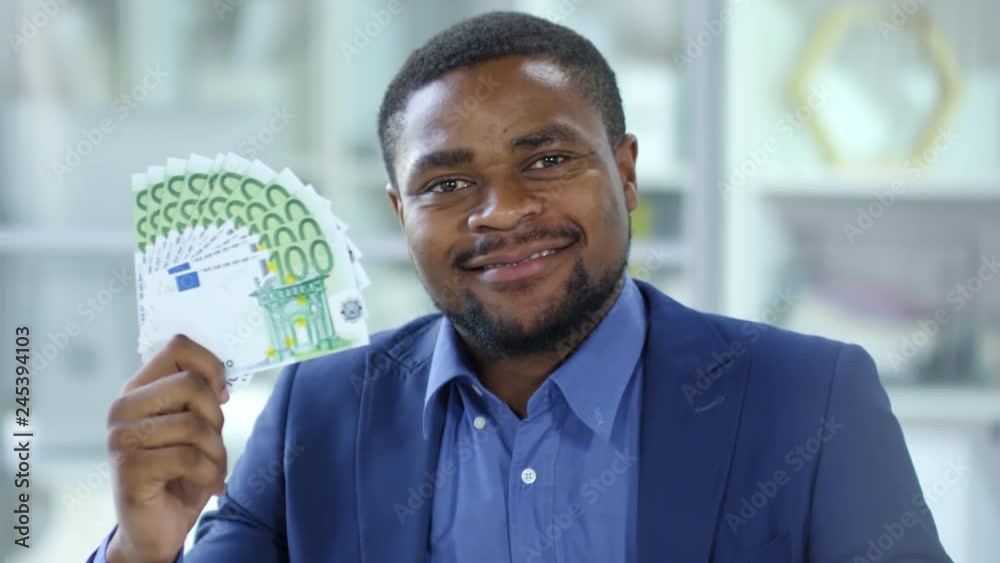 Tracking shot of smiling African American businessman holding euro banknotes fan and looking at camera in the office Tracking shot of smiling African American businessman holding euro banknotes fan and looking at camera in the office