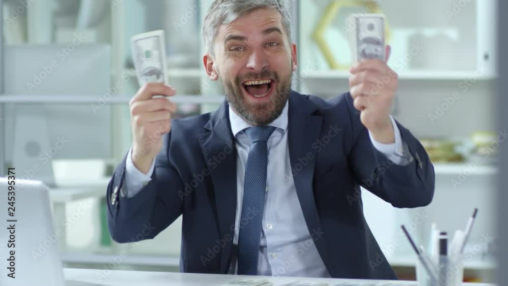 Happy businessman holding dollar banknotes with both hands and shouting something at camera with excitement while sitting at his workplace in the office Happy businessman holding dollar banknotes with both hands and shouting something at camera with excitement while sitting at his workplace in the office
