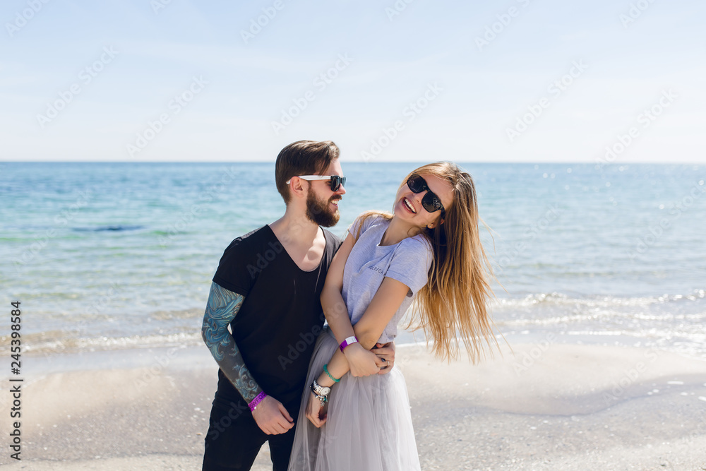 Guy with tattoo on hand is hugging girl with long hair near sea. He is looking at her. She is smiling to the camera. Guy with tattoo on hand is hugging girl with long hair near sea. He is looking at her. She is smiling to the camera.
