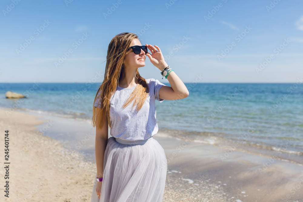 Young pretty girl with long hair is walking on the beach near sea. She wears gray T-shirt with skirt. She is smiling trough black sunglasses to the sun. Young pretty girl with long hair is walking on the beach near sea. She wears gray T-shirt with skirt. She is smiling trough black sunglasses to the sun.