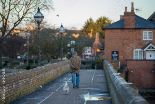lonely man walking across bridge with small white dog with wagging tale following behind