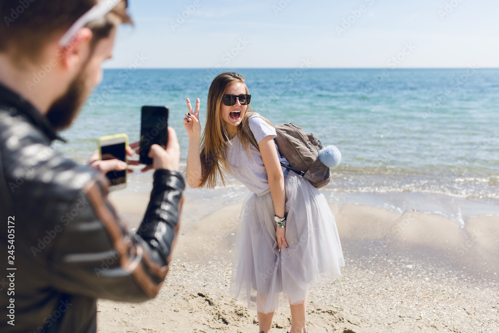 View  of guy from back who photographs the posing girl near sea. She has long hair, gray T-shirt and skirt. She is funny smiling to him.