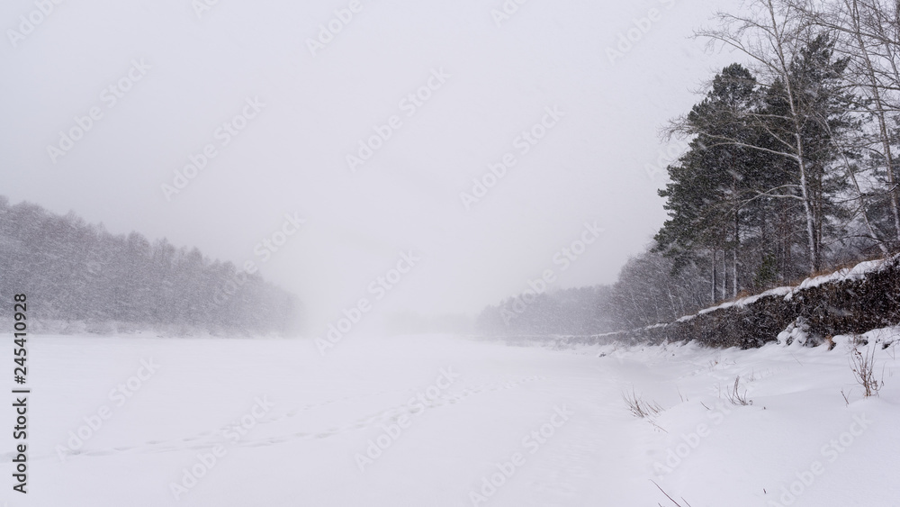 A heavy snowstorm covers the tracks on the ice of the river in winter in the middle of two banks with trees and a cliff. A heavy snowstorm covers the tracks on the ice of the river in winter in the middle of two banks with trees and a cliff.