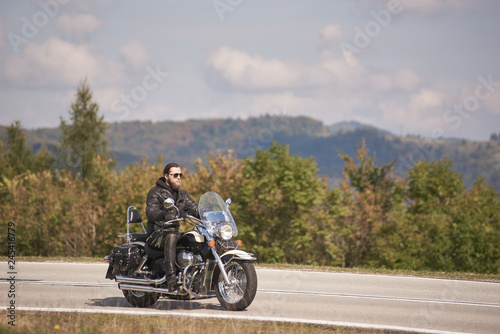 Side view of bearded biker in sunglasses and black leather clothing riding cruiser motorbike along narrow asphalt path on sunny summer day © anatoliy_gleb