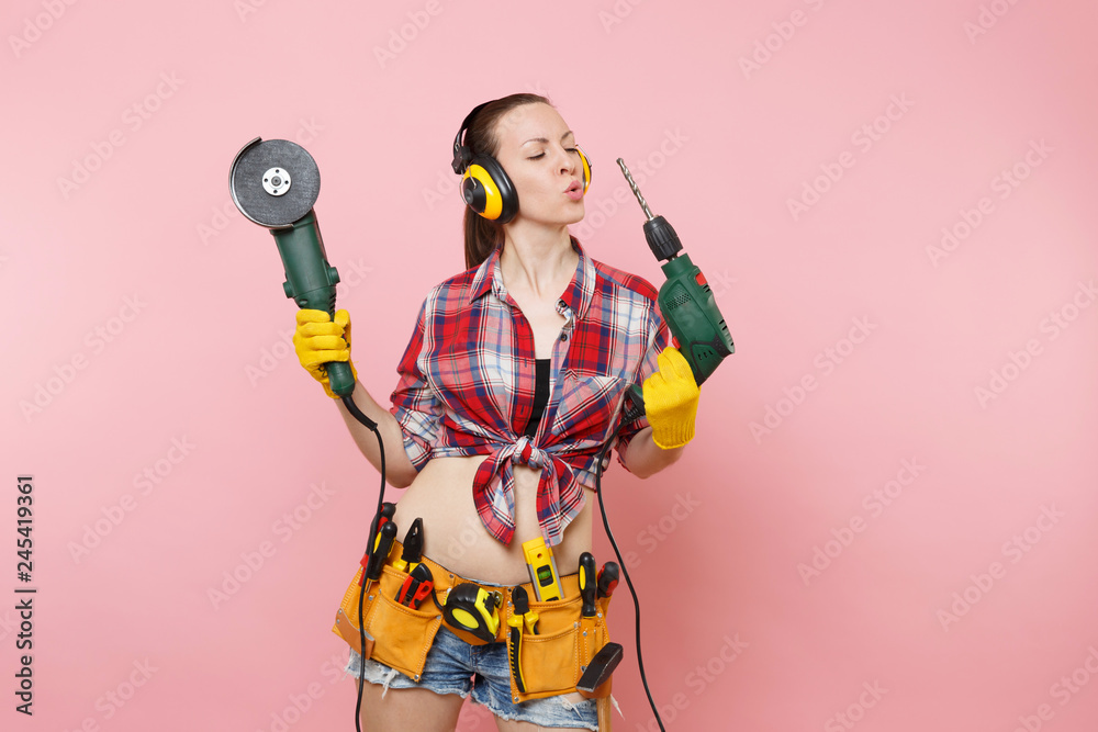 Energy handyman woman in gloves, noise insulated headphones, kit tools belt full of instruments holding power saw electric drill isolated on pink background. Female in male work. Renovation concept.