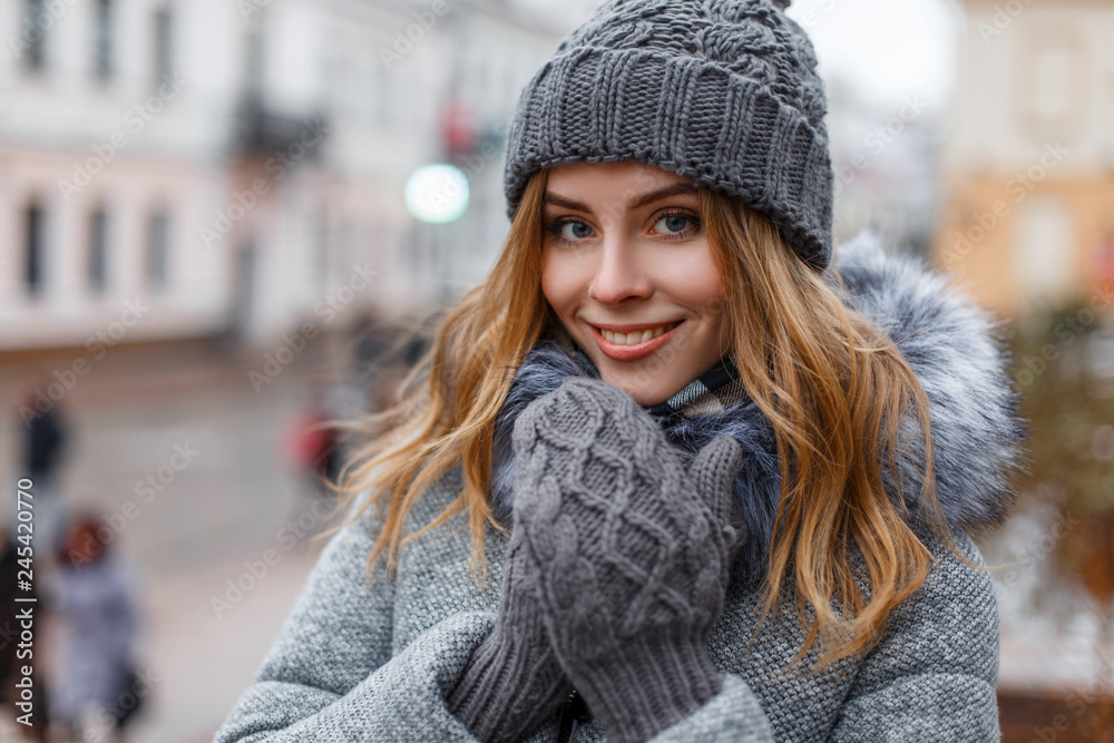 Portrait of a wonderful young woman with beautiful blue eyes with natural make-up in a sweet smile in a knitted hat in knitted mittens in a stylish coat on the background of city streets. Happy girl. - obrazy, fototapety, plakaty 