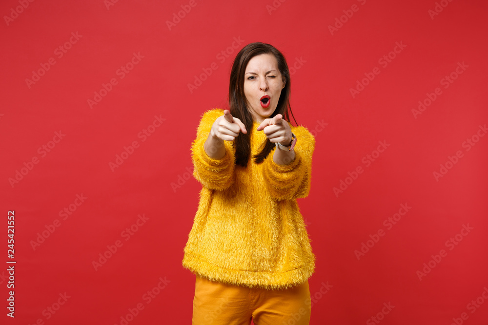 Funny young woman in yellow fur sweater pointing index fingers on camera and blinking isolated on bright red wall background in studio. People sincere emotions, lifestyle concept. Mock up copy space. Funny young woman in yellow fur sweater pointing index fingers on camera and blinking isolated on bright red wall background in studio. People sincere emotions, lifestyle concept. Mock up copy space.