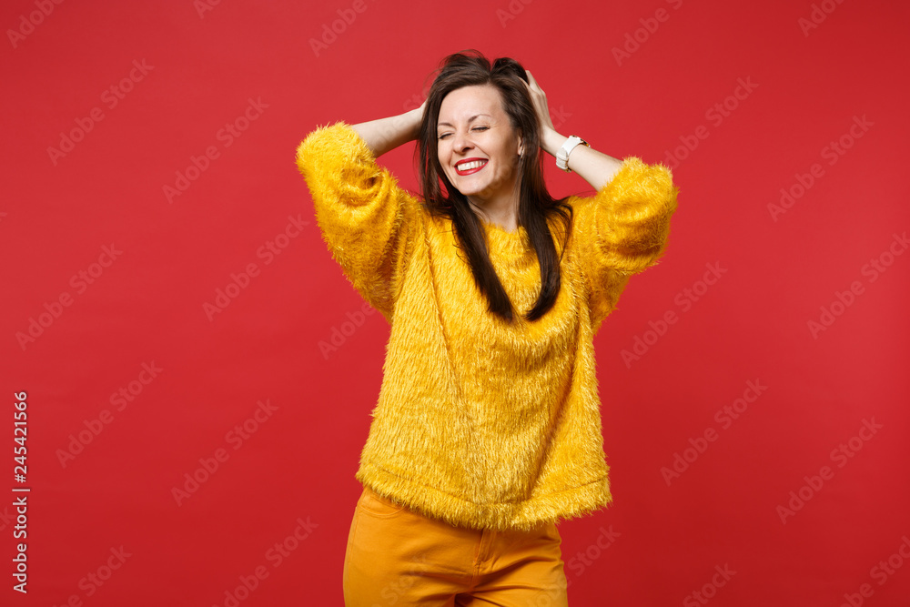 Portrait of smiling young woman in yellow fur sweater keeping eyes closed, clinging to head isolated on bright red background in studio. People sincere emotions, lifestyle concept. Mock up copy space. Portrait of smiling young woman in yellow fur sweater keeping eyes closed, clinging to head isolated on bright red background in studio. People sincere emotions, lifestyle concept. Mock up copy space.