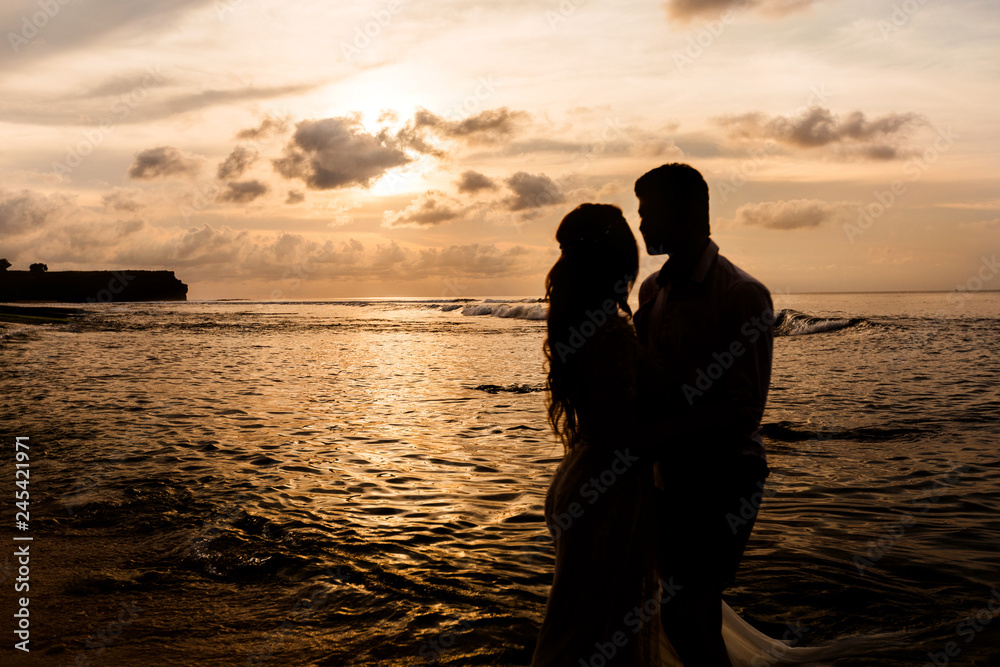 Silhouettes of bride and groom embracing look at each other on the beach at sunset
