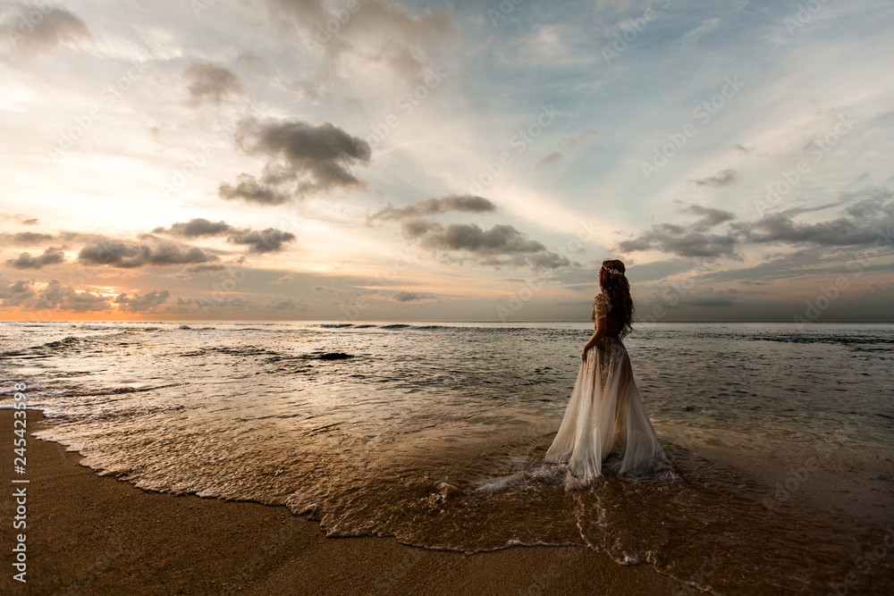 A bride in a white wedding dress on the beach admires the sunset, standing in the sea water.