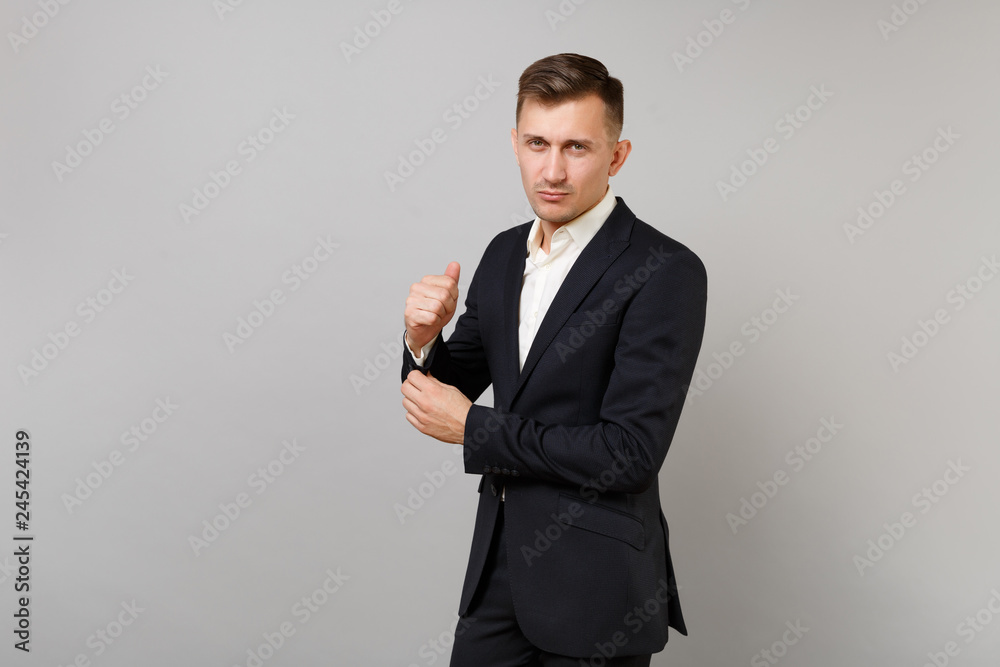 Portrait of handsome young business man in classic black suit, shirt straightening sleeves, cuffs isolated on grey background in studio. Achievement career wealth business concept. Mock up copy space. Portrait of handsome young business man in classic black suit, shirt straightening sleeves, cuffs isolated on grey background in studio. Achievement career wealth business concept. Mock up copy space.
