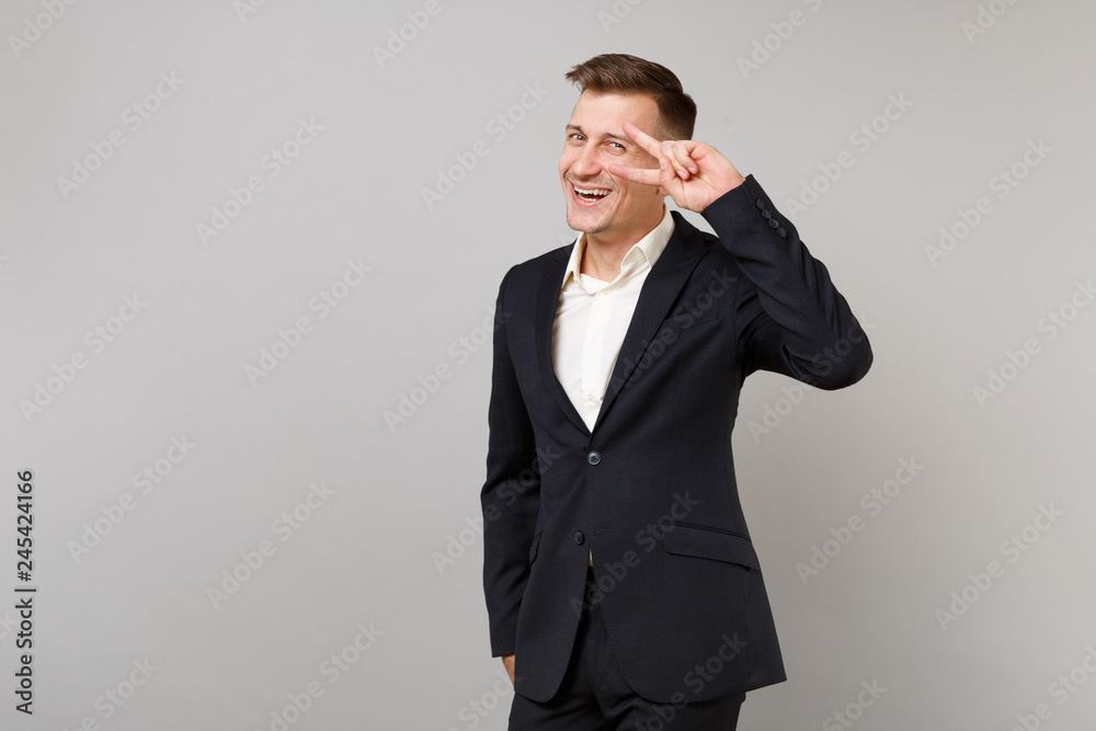 Portrait of joyful young business man in classic black suit white shirt showing victory sign isolated on grey wall background in studio. Achievement career wealth business concept. Mock up copy space. Portrait of joyful young business man in classic black suit white shirt showing victory sign isolated on grey wall background in studio. Achievement career wealth business concept. Mock up copy space.