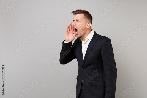 Portrait of irritated young business man in classic black suit, shirt screaming with hand gesture isolated on grey background in studio. Achievement career wealth business concept. Mock up copy space.