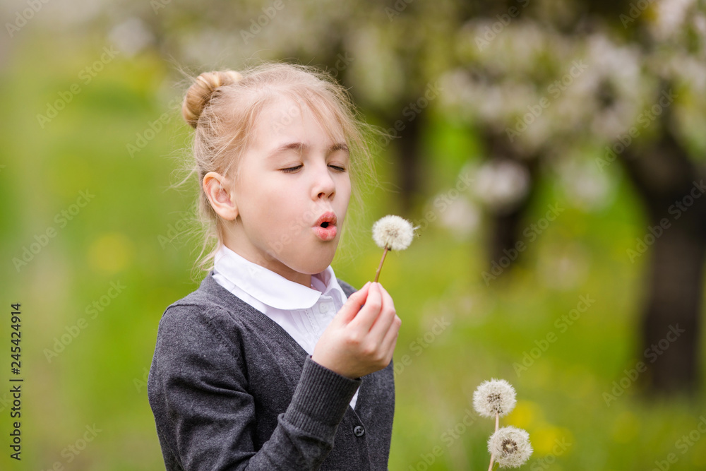 Portrait of a blonde girl in gray jacket standing against background of flowering trees. Kid with white dandellion