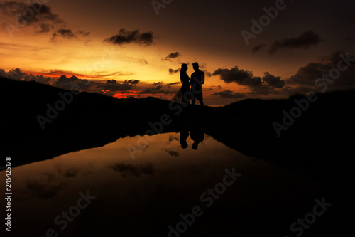 Bride and groom on the beach at sunset, their silhouettes are reflected in the sea water.
