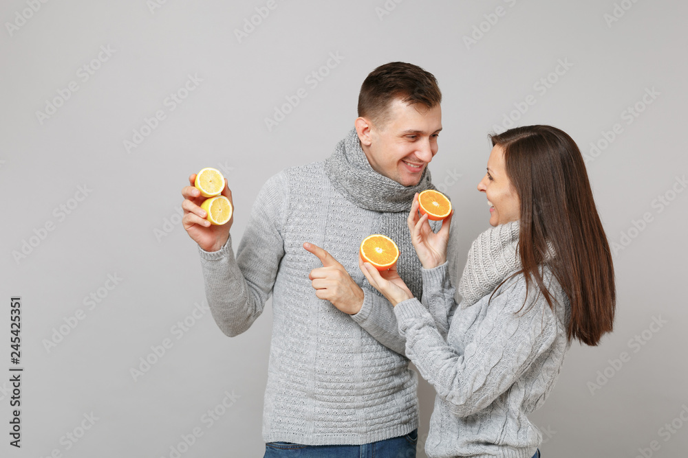 Fun couple girl guy in gray sweaters, scarves together hold orange lemon isolated on grey wall background studio portrait. Healthy lifestyle ill sick treatment cold season concept. Mock up copy space. Fun couple girl guy in gray sweaters, scarves together hold orange lemon isolated on grey wall background studio portrait. Healthy lifestyle ill sick treatment cold season concept. Mock up copy space.