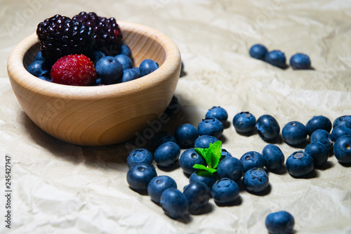 An isolated wooden bowl full of blueberries and wildberries with plain soft background decorated by fresh mint and with a few blueberries around on the paper