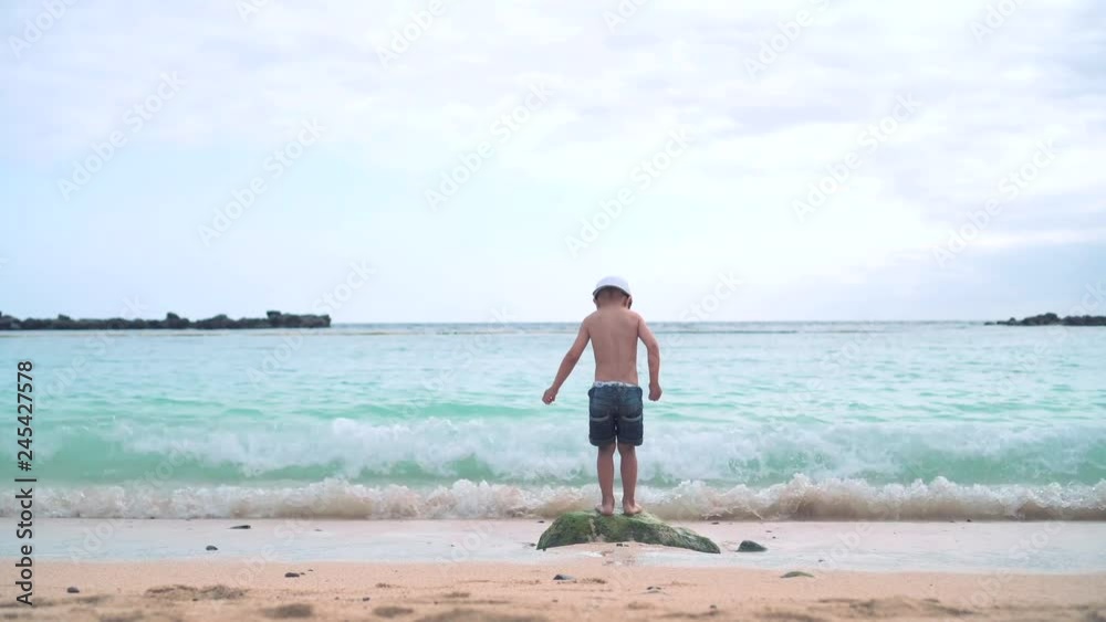 A little boy in shorts, a baseball cap and sunglasses runs up the sandy beach to the stone, jumps on it and plays with the sea wave A little boy in shorts, a baseball cap and sunglasses runs up the sandy beach to the stone, jumps on it and plays with the sea wave