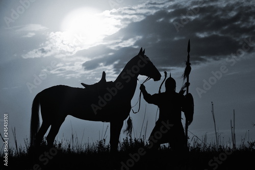 Natural silhouette of asian steppe warrior holding a spear with his battle horse in the sun light behind. Horizontal, black and white.